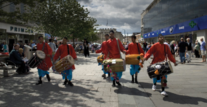 Six bhangra drummers in orange and blue costumes in a line, walking down a high street